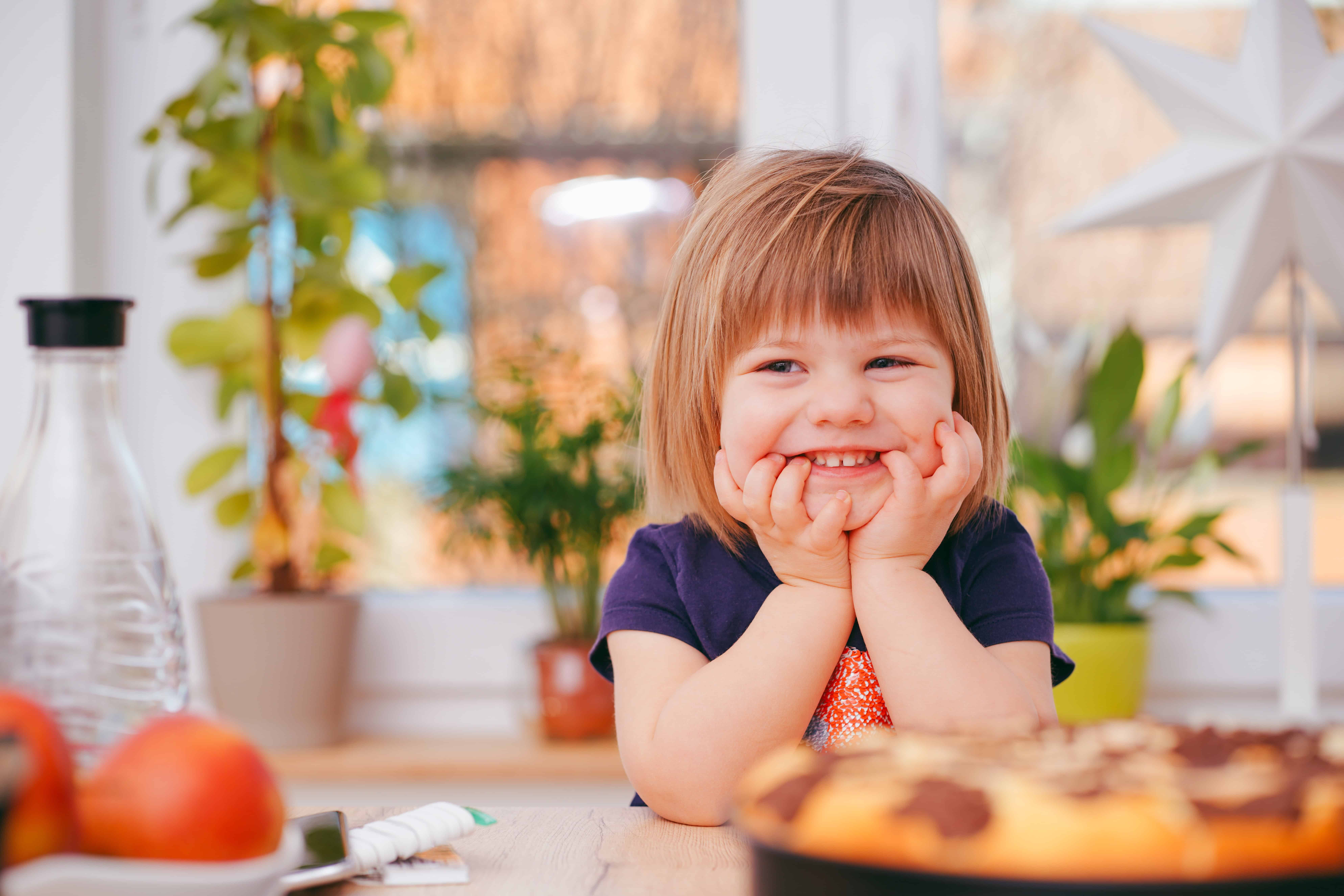 A toddler with brown hair and bangs sits at a table, resting their chin on their hands with a sweet smile during mealtime, illustrating the self-feeding and independence skills that develop during the second year.