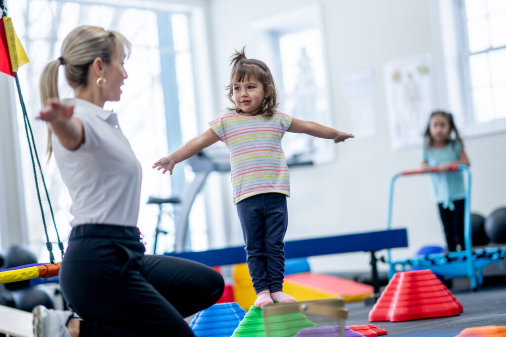 A female Physiotherapist works with a little girl on balance. The little girl is walking on stepping stars while trying to hold her balance at each stop.