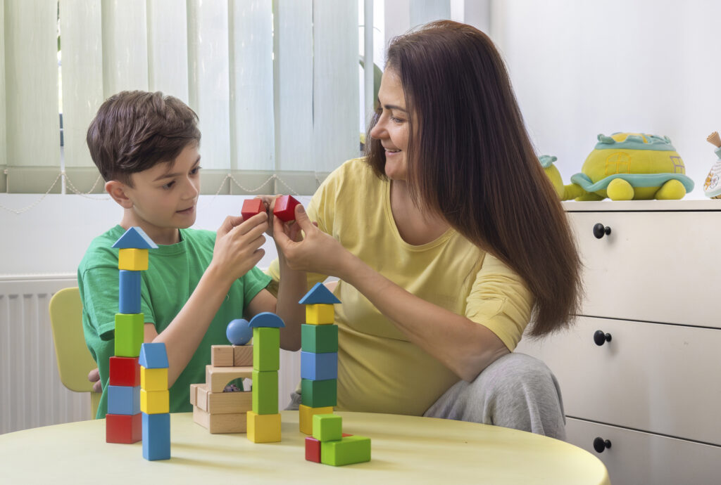 Happy young boy and a woman, possibly his mother or therapist, are playing together with colorful wooden blocks at a table in a bright, cozy home environment. The boy is smiling and engaged, demonstrating focus and curiosity as he explores the tactile shapes and structures. The woman gently supports and encourages him, reinforcing a warm and trusting connection. This scene represents the values of early childhood education, autism therapy, ADHD support, and occupational therapy. It also highlights the importance of sensory learning, emotional bonding, and developmental activities in a nurturing and inclusive setting. Ideal for illustrating topics in child psychology, neurodiversity, home education, and parent-child interaction.