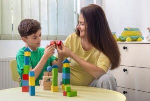 Happy young boy and a woman, possibly his mother or therapist, are playing together with colorful wooden blocks at a table in a bright, cozy home environment. The boy is smiling and engaged, demonstrating focus and curiosity as he explores the tactile shapes and structures. The woman gently supports and encourages him, reinforcing a warm and trusting connection. This scene represents the values of early childhood education, autism therapy, ADHD support, and occupational therapy. It also highlights the importance of sensory learning, emotional bonding, and developmental activities in a nurturing and inclusive setting. Ideal for illustrating topics in child psychology, neurodiversity, home education, and parent-child interaction.